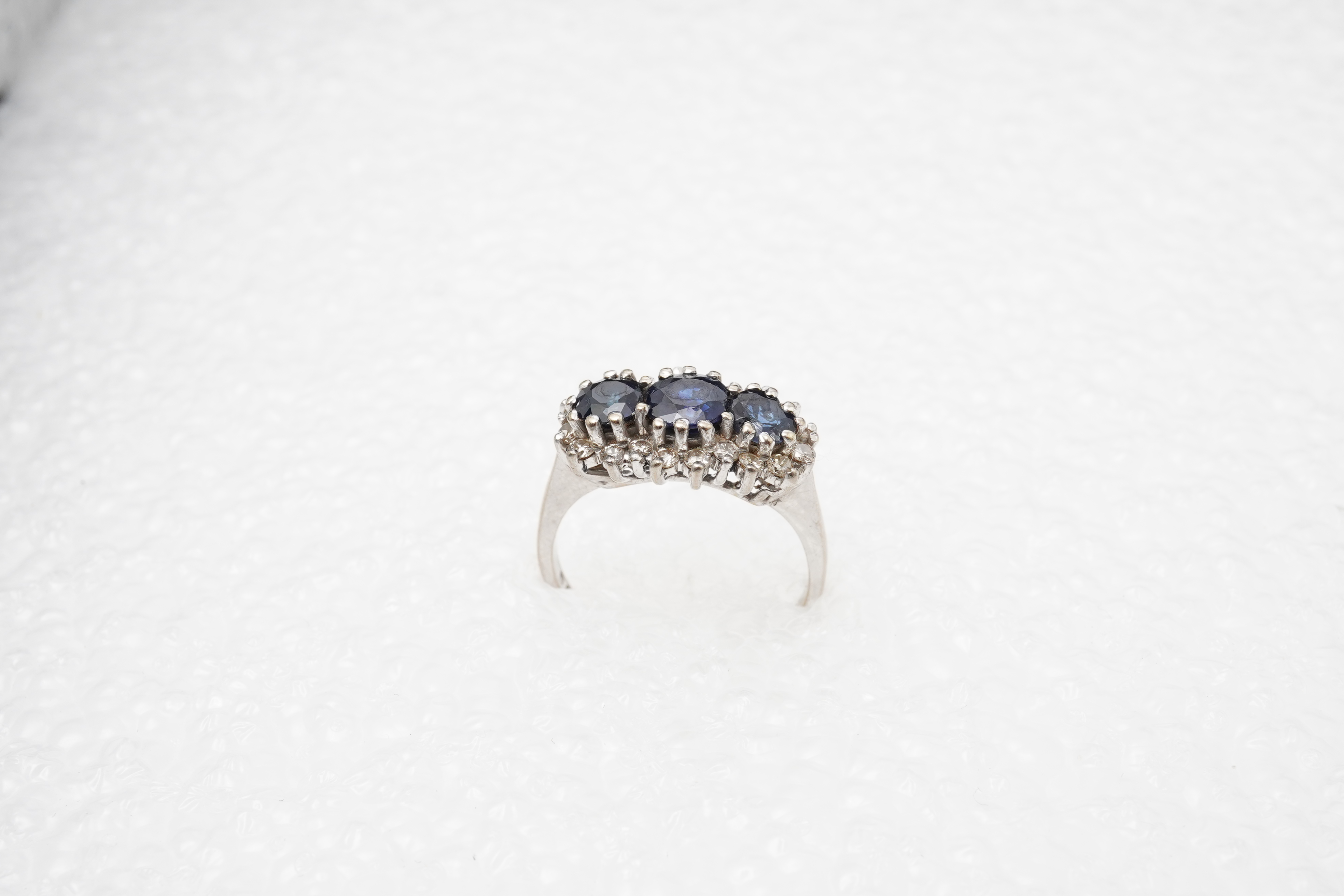 A sapphire and diamond ring, set with three circular-cut and oval sapphires, within a border of brilliant-cut diamonds totalling approximately 0.40 carats, mounted in 18ct white gold, size M1/2, British hallmarks, gross